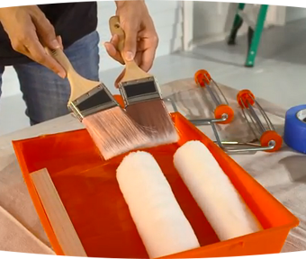 Person holding a paint brush in each hand and two paint rollers in a tray