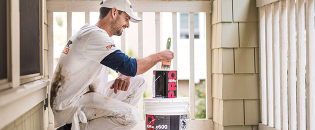 Pro Contractor wearing a cap and shirt with Behr logo dipping a paint brush on a 1 gallon of BEHR PRO e600 paint can on a porch that he is going to paint.