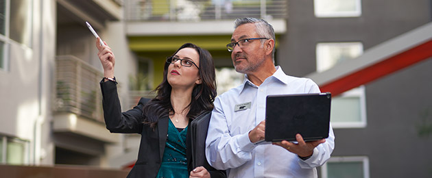 A Large Image of a Property Manager showing a BEHR PRO Rep a multi-family complex. The property manager is pointing at something while the BEHR PRO Rep is looking at what she is pointing and taking notes on a laptop.