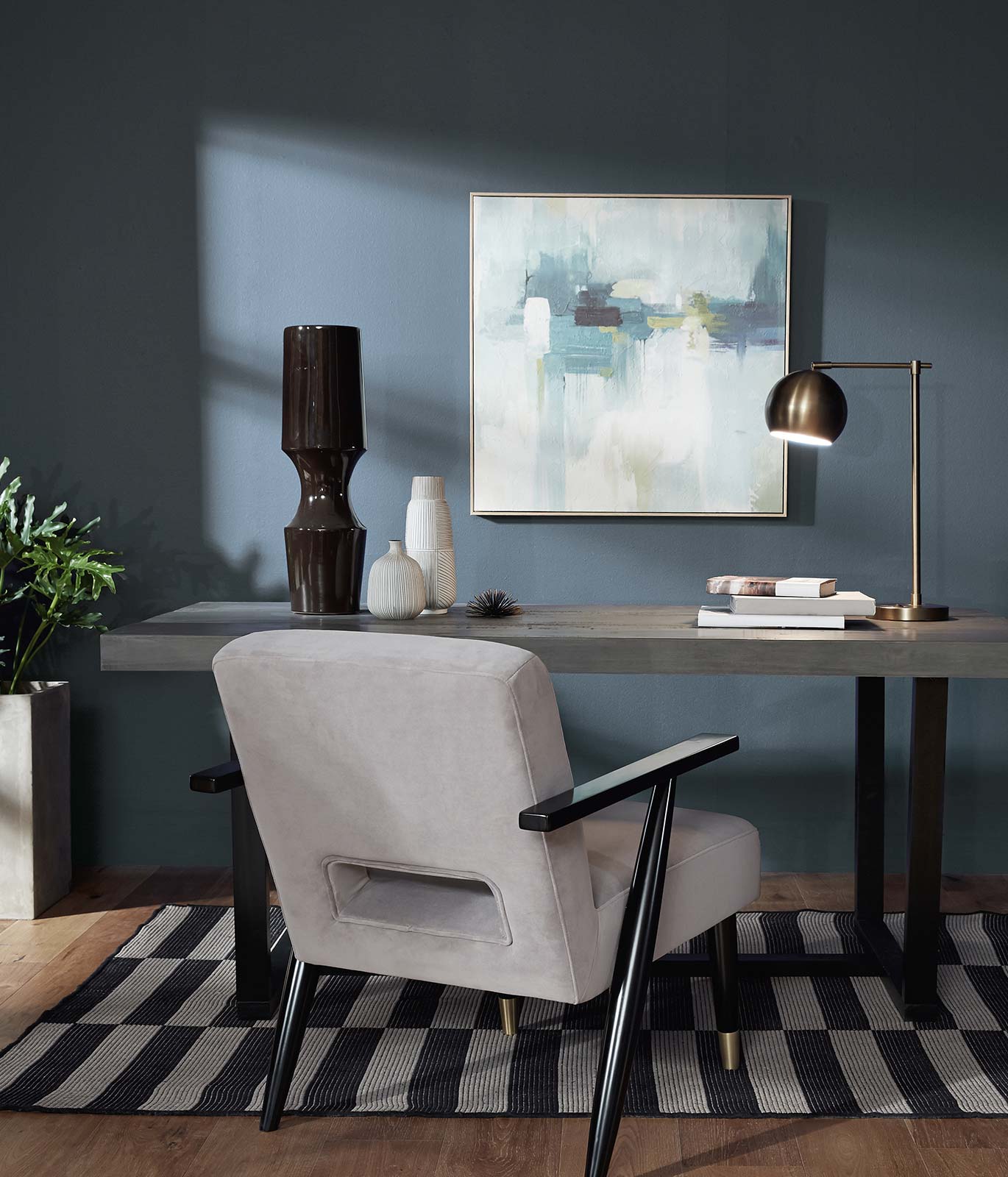 An office with a gray colored wood desk sitting against a wall painted in blue. There is a abstract painting hanging on the wall. The mood is relaxed and calm.