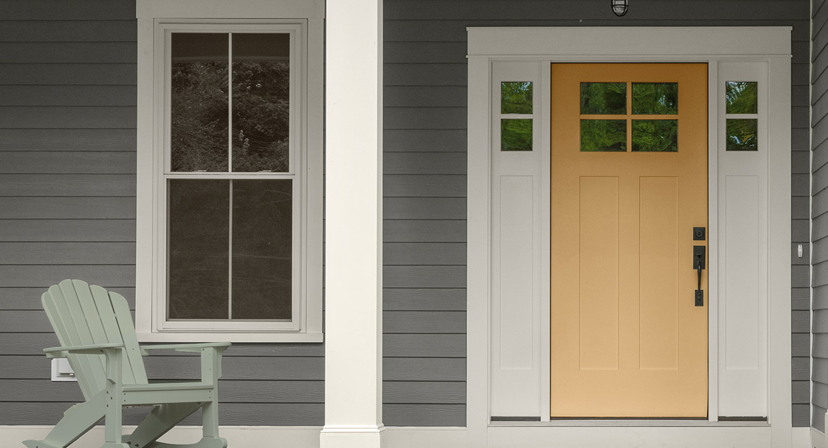 The front porch of a gray painted home with a yellow door and a green rocking chair. The mood is relaxed.