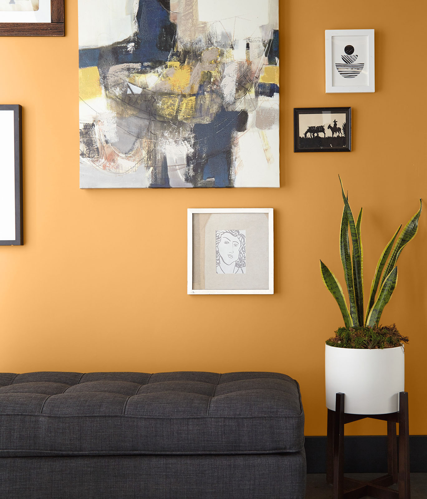 A living room painted in golden-yellow with a gray couch. The wall is filled with hanging art work. The room feels optimistic and bright. 