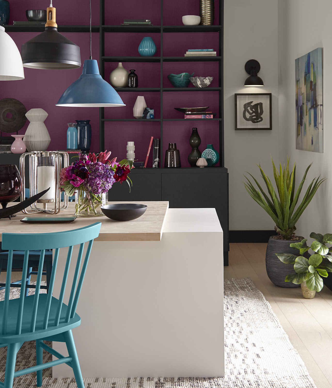 A dining room with a white and wood table in the foreground. In the background is a bookshelf painted a magenta color inside the shelves and black for the cabinet. The room gives off an optimistic vibe.